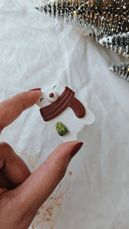 Hand holding a small snowman ornament with a brown scarf and green hat on a white background.