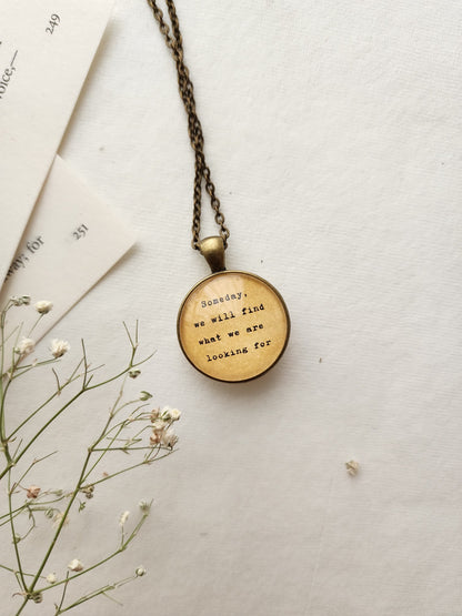 A brass pendant necklace with a quote inscribed, displayed on a neutral background with some floral elements.